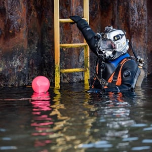 Berufstaucher in voller Ausrüstung hält sich an einer gelben Leiter an einer rostigen Kaimauer fest, neben einer roten Markierungsboje im Wasser.