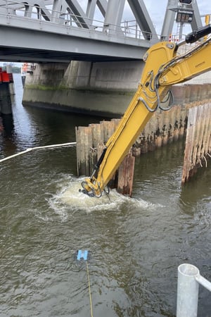 Unterwasser-Rückbau an der ehemaligen Rethebrücke in Hamburg: Baggerarm mit hydraulischem Werkzeug arbeitet im Spundwandfeld; Arbeiten begleitet von Kampfmittelsondierung und Taucher.